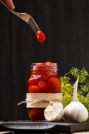 Photo for Pickled cherry tomatoes in an open transparent jar, one canned tomato on a vintage fork over a jar, garlic, dill on a dark wooden background. home preservation. - Royalty Free Image