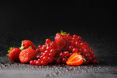 Foto de Strawberries and red currants on a dark background. Sprigs of red currant and strawberries with green tails on a black background with water drops - Imagen libre de derechos