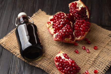 Ripe broken pomegranate, scattered large grains, freshly squeezed pomegranate juice in a beautiful bottle on a wooden and textile background. Creative composition of pomegranate and pomegranate juice.