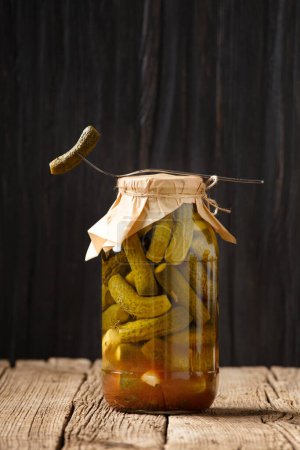 Photo for Homemade pickled cucumbers in a transparent glass jar with a craft lid, one cucumber on a fork lies on top. Canned gherkins on a dark wooden background with space for text. - Royalty Free Image