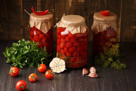 Foto de Marinated cherry tomatoes with garlic, dill and spices. Homemade canned tomatoes in closed jars with craft lids, fresh hot peppers and tomatoes on a wooden background. Pickled vegetables. - Imagen libre de derechos