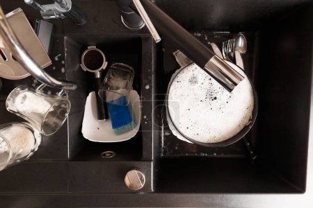 Foto de Concepto de lavar platos. Platos sucios con espuma de detergente en un fregadero de piedra negra en la vista superior de la cocina. Vasos sucios, tazas, platos, cubiertos en el fregadero de la cocina. - Imagen libre de derechos