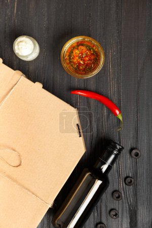 Photo for Pizza in a closed box, sauces and vegetables on a dark wooden background top view with space for text or advertising. Food delivery. - Royalty Free Image