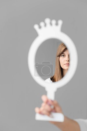 Foto de La mano de una mujer sostiene un espejo de mesa, que refleja el rostro de una joven y hermosa niña. Reflejo fotográfico conceptual de un rostro femenino joven en un espejo sobre un fondo claro. - Imagen libre de derechos
