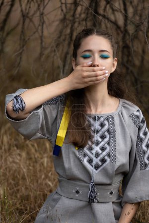 Téléchargez les photos : Une jeune fille au ruban bleu et jaune se couvrait la bouche de sa main. La douleur de l'Ukraine. Symbole du peuple ukrainien. Guerre en Ukraine. La Russie se moque et tue le peuple ukrainien. Génocide en Ukraine - en image libre de droit