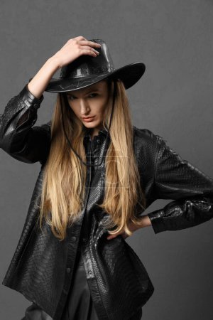 Téléchargez les photos : Jolie jeune fille sexy sensuelle à la mode posant magnifiquement sur un fond sombre dans une chemise en cuir noir et un chapeau de cow-boy, publicité de vêtements, une femme dans un chapeau en cuir noir. - en image libre de droit