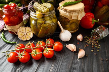 Photo for Canned cucumbers and tomatoes in jars, seasonings and spices, fresh cherry tomatoes and peppers on a dark wooden background. The concept of home canning. - Royalty Free Image