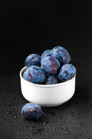 Téléchargez les photos : Une poignée de prunes bleues dans une assiette blanche sur un fond sombre. - en image libre de droit