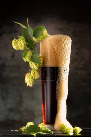 Téléchargez les photos : Bière foncée dans une tasse et houblon vert sur table en bois sur fond brun - en image libre de droit