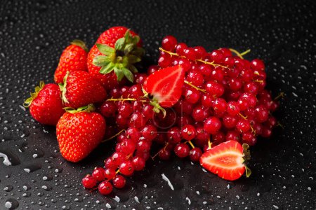 Foto de Strawberries and red currants on a dark background. Sprigs of red currant and strawberries with green tails on a black background with water drops - Imagen libre de derechos