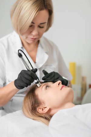 Foto de Rejuvenating facial therapy treatment at spa salon clinic. Young beautiful woman getting lifting anti-aging, face massage and skincare by electroporation facial therapy aesthetic cosmetology. - Imagen libre de derechos