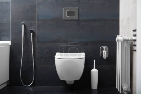 Photo for Modern wall-mounted white toilet bowl, chrome flush button and bidet hygienic shower against the background of a black bathroom wall. Part of the interior of the bathroom in the apartment. - Royalty Free Image