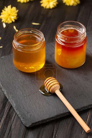 Photo for Honey in jars, dipper and flowers on a black stone plate on a wooden background. The concept of organic bee products. - Royalty Free Image
