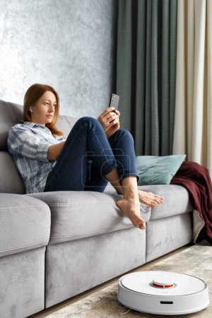 Foto de Una chica se sienta en un sofá en la sala de estar y controla una aspiradora robótica con un teléfono inteligente. Casa Inteligente. El concepto de electrodomésticos inteligentes, tareas domésticas y tecnología. - Imagen libre de derechos