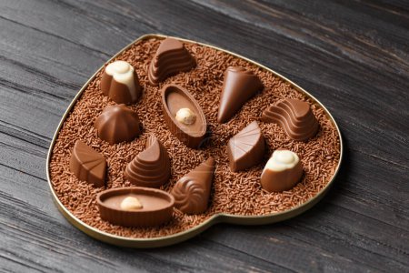 Foto de Dulces de chocolate en chispas de chocolate en una caja en forma de corazón sobre un fondo de madera oscura de cerca, el Día de San Valentín. - Imagen libre de derechos