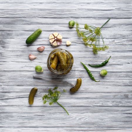 Photo for Pickled cucumbers in an open jar, gherkin on a fork and garlic on a light wooden background, top view, copy space. Homemade canned cucumbers. - Royalty Free Image