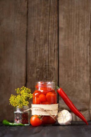 Photo for Pickled cherry tomatoes in an open glass jar, chili peppers, garlic, spices and herbs for marinade on a dark wooden background. Home preservation. - Royalty Free Image