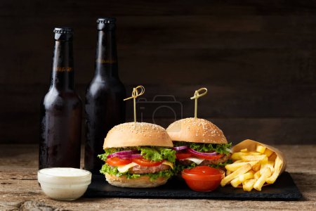 Foto de Dos hamburguesas, papas fritas, cerveza en botellas oscuras sobre un fondo de madera. Comida rápida, hamburguesas caseras - Imagen libre de derechos