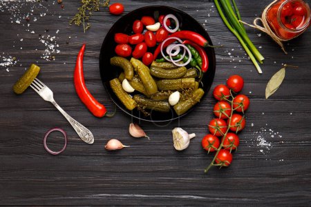Photo for Homemade canned tomatoes, cucumbers, chili peppers in a round black plate, gherkin on a vintage fork on a dark wooden background. - Royalty Free Image