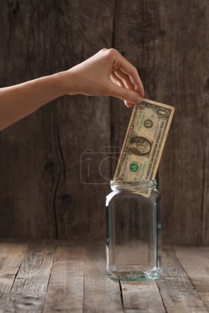 Téléchargez les photos : Une main met un dollar de papier dans un bocal en verre sur un fond en bois. Le concept d'accumulation, de stockage, de sécurité, d'épargne, de méfiance envers les banques. - en image libre de droit