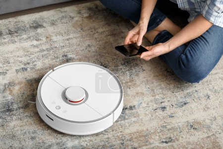 Photo for A girl sits on the floor in the living room and controls a robotic vacuum cleaner using a smartphone. Smart House. The concept of smart home appliances, housework and technology. - Royalty Free Image