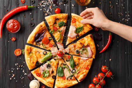 Foto de Hands take slices of cut pizza on a dark wooden background top view. Pizza with salami, jerky, sauces, spices, fresh cherry tomatoes on a black background. Women's hands share a pizza. - Imagen libre de derechos