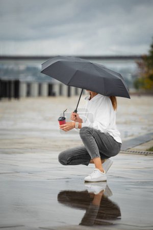 Foto de Una chica bajo un paraguas en tiempo lluvioso camina por la calle y bebe café. - Imagen libre de derechos