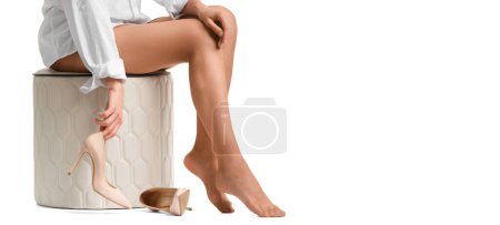 Téléchargez les photos : Une jeune femme enlève ses chaussures à talons hauts et les tient dans ses mains alors qu'elle est assise sur un pouf, en gros plan. Jambes fatiguées, douleurs aux jambes, douleurs articulaires, chaussures inconfortables. - en image libre de droit
