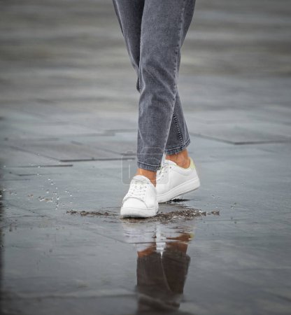 Téléchargez les photos : Jambes d'une fille en jean et baskets blanches marchent par temps pluvieux dans une rue de la ville, gouttes de pluie, éclaboussures d'eau, réflexion dans les flaques d'eau - en image libre de droit