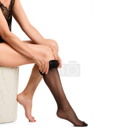 Photo for A young slender girl puts on a black stocking on her leg, sitting on a pouffe on a white background. - Royalty Free Image