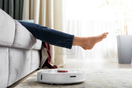 Photo for The robot vacuum cleaner does the cleaning in the living room. The girl raised her legs to let the robotic vacuum cleaner pass. The concept of smart home, technology, housework. - Royalty Free Image