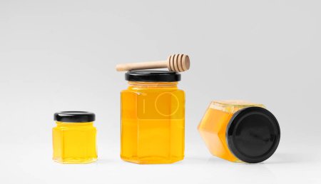 Foto de Composición creativa de frascos de vidrio en forma de panales de diferentes tamaños con miel sobre un fondo blanco de cerca. Frascos de vidrio con miel orgánica en diferentes posiciones y dipper en aislamiento. - Imagen libre de derechos