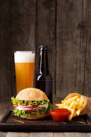 Foto de Hamburguesa, cerveza en una botella y en una taza de vidrio, patatas fritas, salsa sobre un fondo de madera, concepto de comida rápida. - Imagen libre de derechos