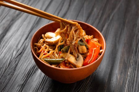 Photo for Plate with buckwheat noodles with vegetables, mushrooms, chicken meat on a dark background. Chinese sticks take Japanese soba from a clay bowl on a wooden background. - Royalty Free Image