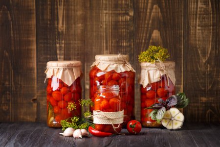 Foto de Tomates cherry enlatados en frascos cerrados y abiertos, especias y hierbas para marinar sobre un fondo de madera, espacio para copiar. - Imagen libre de derechos
