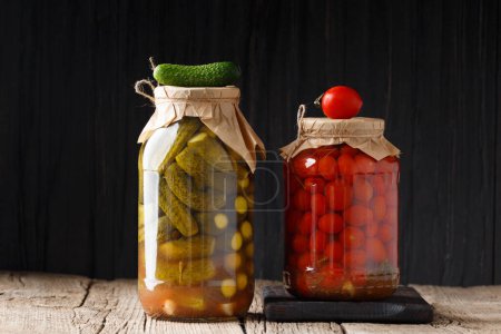 Photo for Canned cucumbers and cherry tomatoes in closed glass jars on a dark wooden background, fresh gherkin and tomato lie on top of the lids. - Royalty Free Image