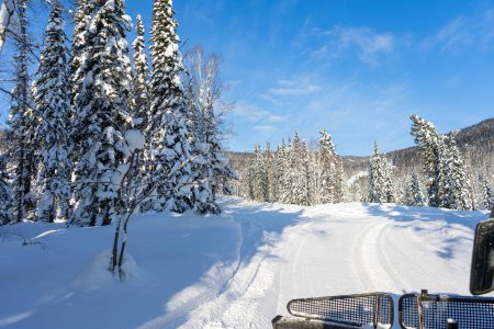 Winter road. Red snowcat rides in a snowy spruce forest, view from the cab. Freeride trip for skiers and snowboarders. Blue sky. Winter activities, adventures, travel. High quality photo