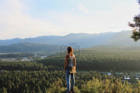 Concept woman traveler on a solo journey, standing on a hill, looking into the distance. Summer photo at sunset, green forest in warm weather. Nature landscape. Hiking outdoor