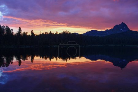 Incroyablement beau coucher de soleil sur un lac de montagne. Des silhouettes d'arbres et un pic rocheux se reflètent dans l'eau. Ciel violet et traînée de soleil jaune. Parc naturel d'Ergaki, région de Krasnoïarsk, Sibérie