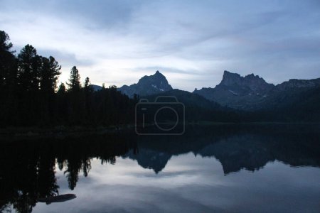 Silhouettes of two rocky mountain peaks and pine trees are reflected in the water of the Svetloye lake. Nature landscape in twilight in Ergaki nature park, Krasnoyarsk region, Siberia, Russia