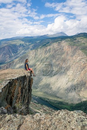 Frau sitzt am Rand einer steilen Klippe über einer tiefen Klippe, die Beine baumeln herunter. Tschulyshman-Tal, Katu-Jaryk-Pass, Altai. Reisen Sie um die Welt. Vertikales Foto