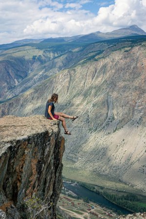 Dangerous photos in nature. A woman sits on the edge of a sheer cliff above a high cliff and dangles her legs. fatal risk for photography. Summer Altai, Chulyshman valley