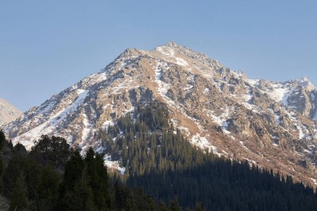 Foto de Alta montaña rocosa con nieve, iluminada por el cálido sol poniente. Bosque de coníferas a la sombra. Paisaje natural en el Parque Nacional Ala-Archa en Kirguistán. - Imagen libre de derechos