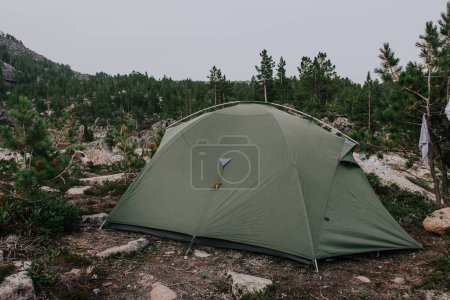 Téléchargez les photos : Camping en forêt lors de randonnées en montagne. Une tente touristique verte pour trois personnes installée dans une forêt entourée de conifères et de rochers à feuilles persistantes dans une zone montagneuse. Équipement dont un voyageur a besoin - en image libre de droit