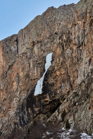 Cascada congelada en una roca de alto acantilado de color marrón. Se derrumbó Icefall en las montañas. Paisaje natural de primavera temprana. El agua se congeló en hielo.