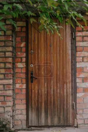 Brown wooden old door made from slats in Red brick house surrounded by green tree foliage. Entrance with two keyholes, peephole and black handle