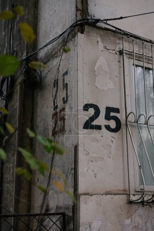 Die Zahl 25 wird mit einer Schablone dreimal auf eine graue Wand an der Ecke des Hauses gemalt, in der Nähe von Fenstern mit Gittern. Hausnummerierung 25. 