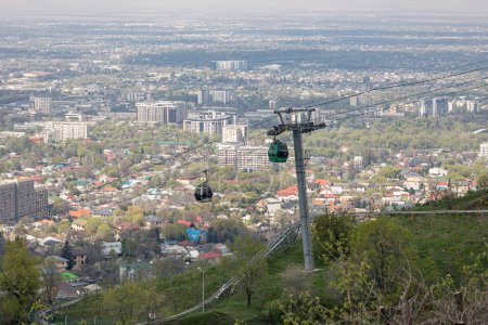 Seilbahn mit zwei Kabinen, vor der Kulisse der Stadt zur Frühlingszeit. Seilbahn auf den Kok-Tobe-Hügel in Almaty, Kasachstan. Touristischer Ort, Wahrzeichen der Stadt. Stützpfeiler der Seilbahn