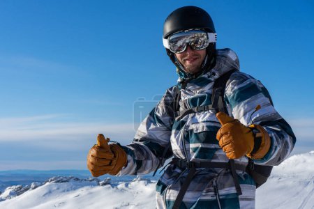 Happy smiling man in skiing or snowboarding winter equipment shows thumbs up, recommendation right choice. Warm jacket, brown gloves, white ski goggles, black sport helmet. Activities at sunny day 