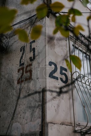 Hausnummerierung 25. Die Zahl 25 wird mit einer Schablone dreimal auf eine graue Wand an der Ecke des Hauses gemalt, in der Nähe eines Fensters mit Gittern. 